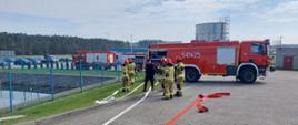 Strażacy w ubraniach specjalnych w trakcie rozwijania węży pożarniczych. Po lewej stronie zbiornik przeciwpożarowy, w tle wozy strażackie ustawione bokiem na terenie zakładu.