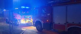 Na zdjęciu dwa pojazdy gaśnicze. Kolor czerwony, włączona sygnalizacja ostrzegawcza. Światła mijania włączone w jednym z pojazdów. Pora nocna. Na pierwszym planie chodnik, znak drogowy, trawnik.