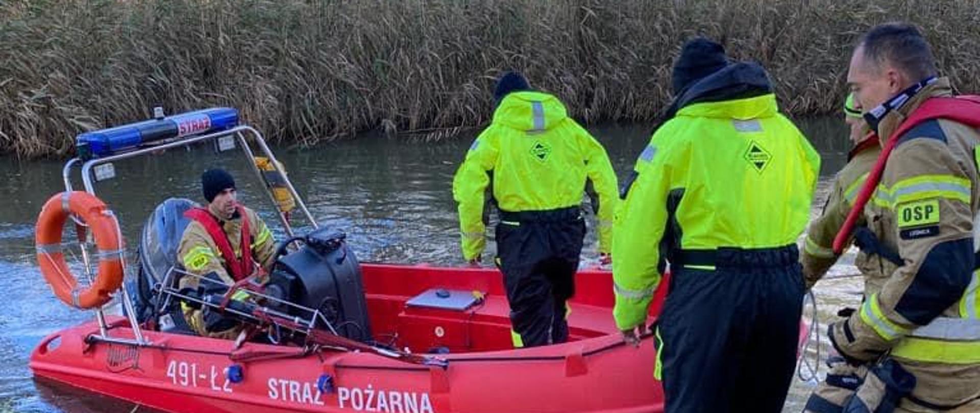Na zdjęciu znajduje się łódź, do której wchodzą strażacy, w tle widać drzewa i domy