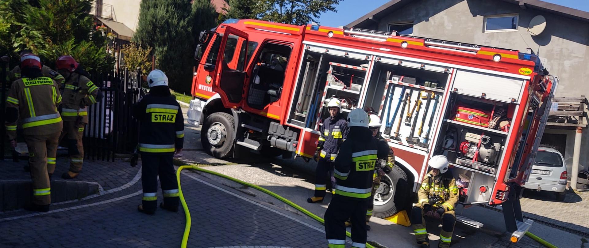 Strażacy oraz \rozwinięte linie wężowe obok samochodu pożarniczego.
