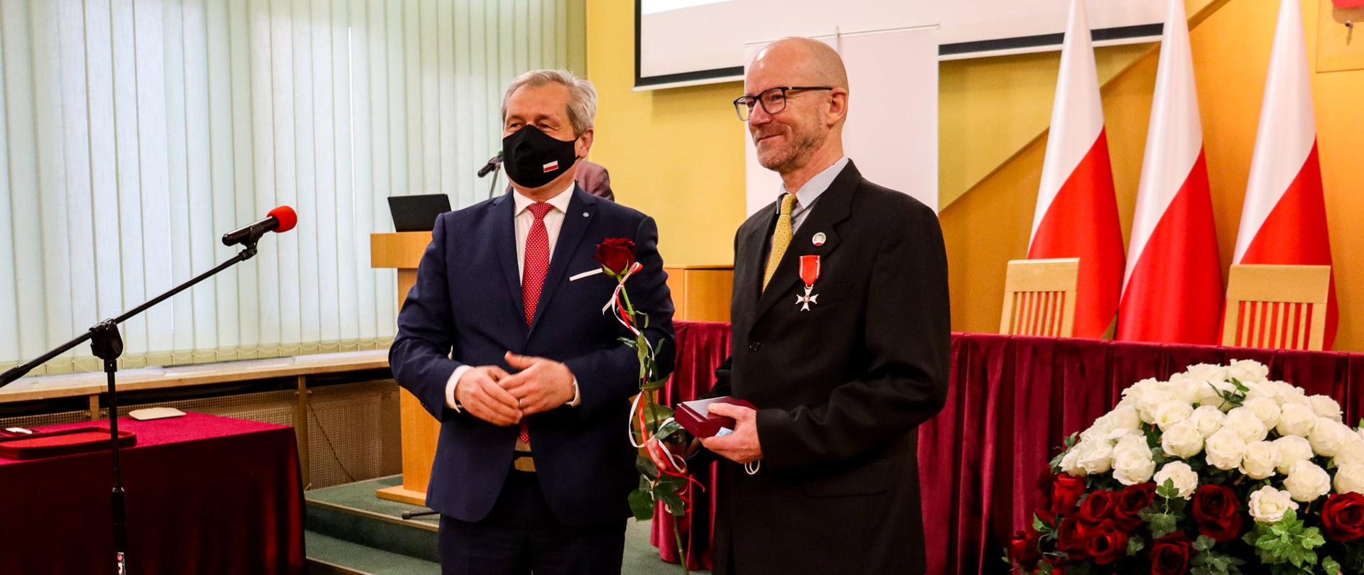 Na zdjęciu wojewoda opolski, stojący obok mężczyzny, który w dłoni trzyma róże oraz bordowe etui na odznaczenie państwowe. Mężczyzna stojący obok wojewody jest osobą wyróżnioną Krzyżem Kawalerskim Odrodzenia Polski. Krzyż zdobi jego ciemny garnitur. W tle widać mównice, biało-czerwone kwiaty. 