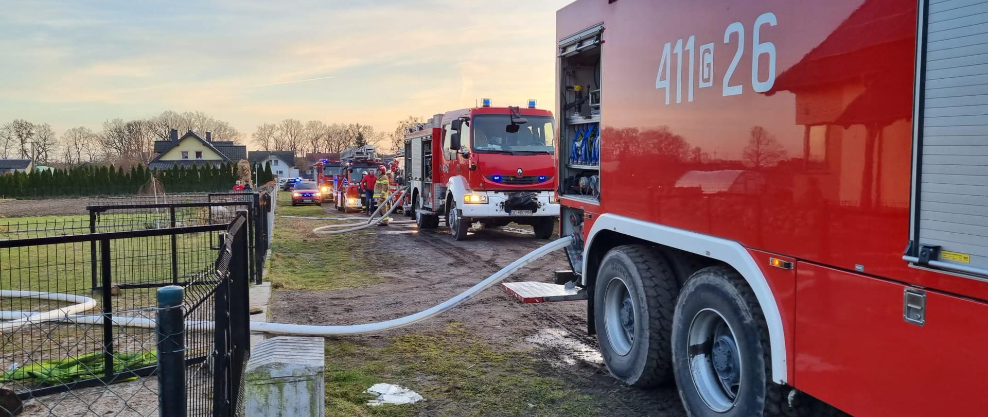 Na drodze gruntowej wzdłuż ogrodzenia stoją samochody strażackie z rozwiniętą linią gaśniczą. W tle zabudowania.