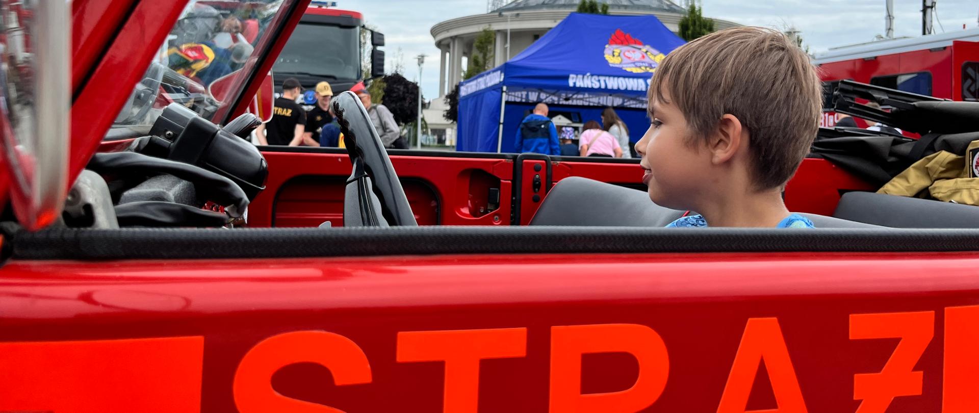 Chłopiec w strażackim samochodzie, za nim widać kościół oraz stoiska promocyjne PSP