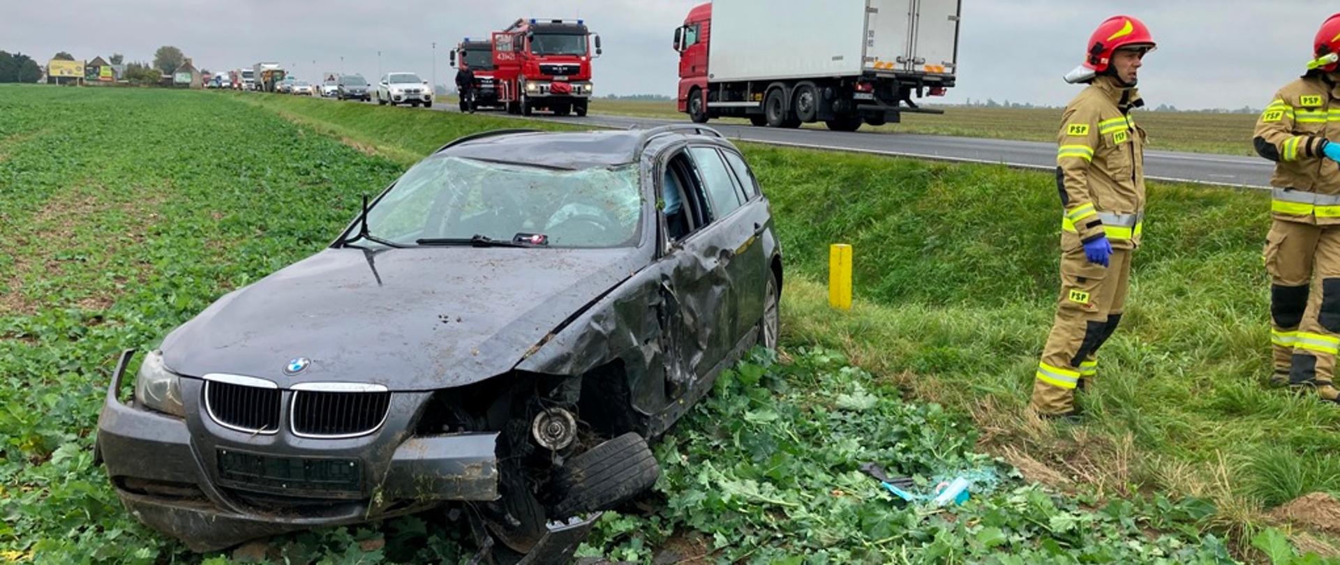 Zdjęcie przedstawia uszkodzony samochód osobowy na polu, który uległ wypadkowi. Obok stoją strażacy.