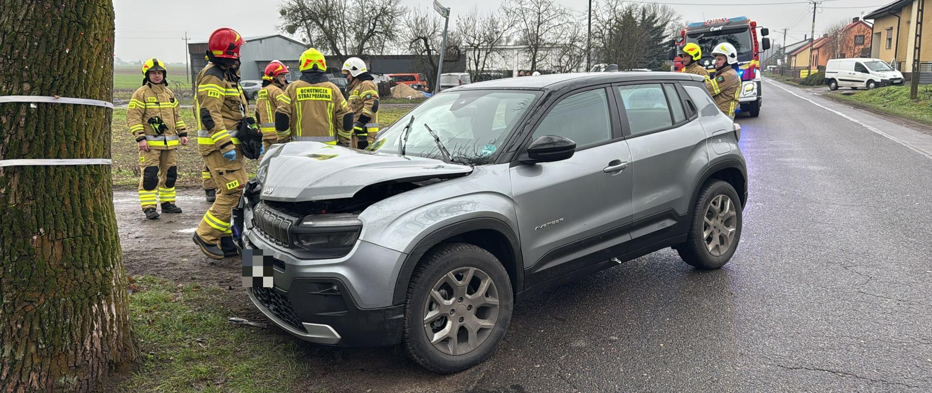 Uszkodzony samochód po uderzeniu w drzewo za nim strażacy w oddali samochód strażacki