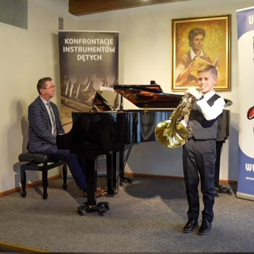Uczeń gra na waltorni stojąc przed fortepianem, mężczyzna gra na fortepianie, w tle baner Konfrontacje Instrumentów Dętych.