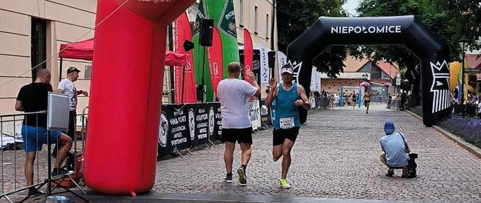 mł.asp. Michał Szewczyk pokonując metę przebiega pod czerwonym balonem w kształcie ramy. mężczyzna w sportowym stroju biegnie po ulicy z kostki brukowej. 