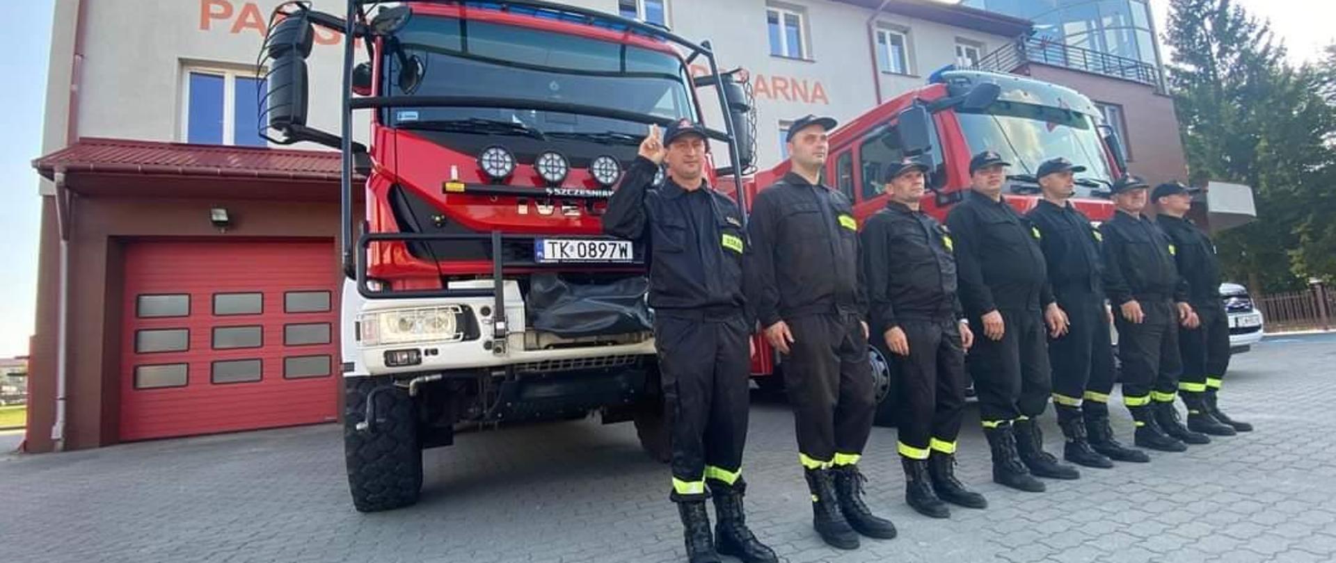 Przed budynkiem KP PSP we Włoszczowie, strażacy podziału bojowego w ubraniach koszarowych, ustawieni w szeregu, pojazdy pożarniczej mają włączone sygnały dźwiękowe i świetlne