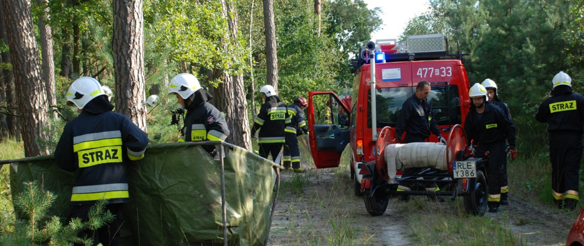 Na zdjęciu widzimy dwóch strażaków przenoszących pusty zbiornik brezentowy, trzech strażaków dopinających wózek wężowy od busa strażackiego oraz dwóch strażaków rozwijających linie wężową. Wszystko odbywa się w scenerii drogi leśnej i lasu.
