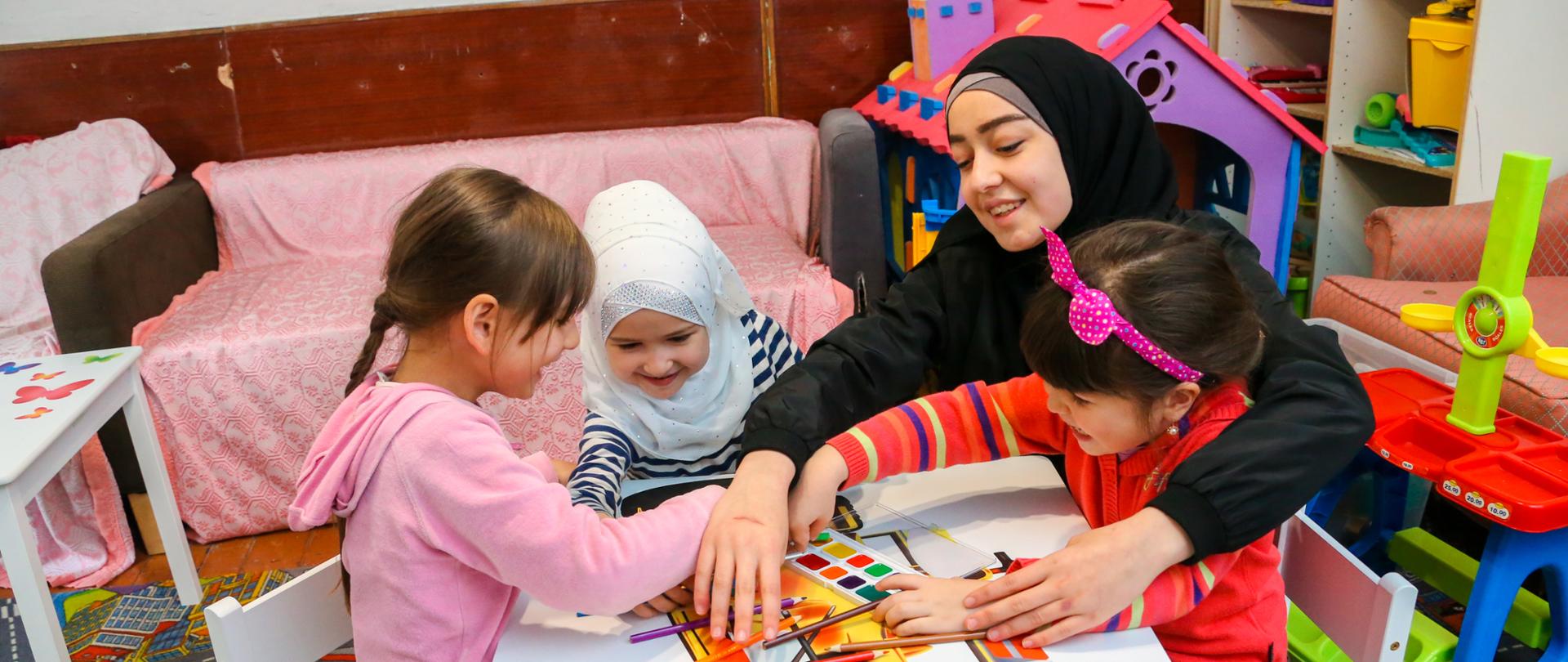 uśmiechnięta kobieta siedzi przy stole z trzema dziewczynkami i rysują na stoliku 