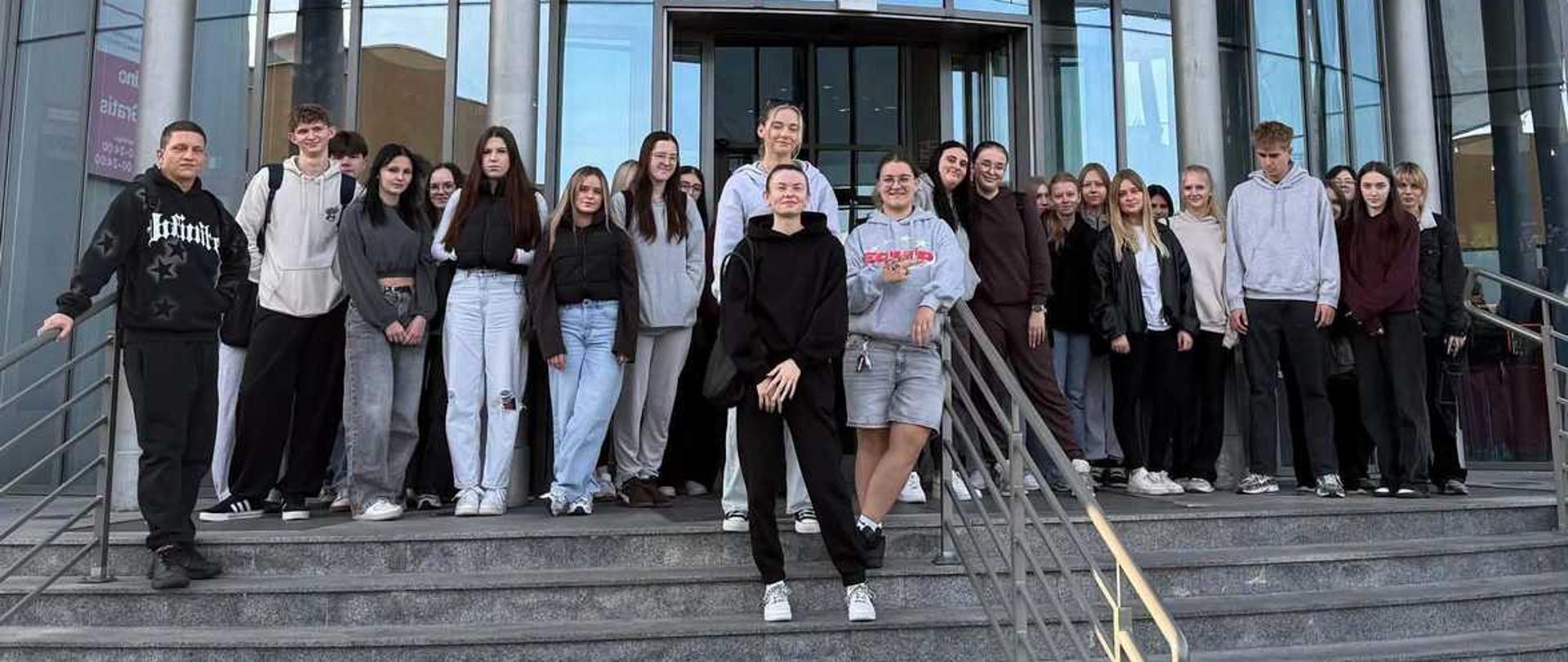 "Zdjęcie przedstawia grupę około dwudziestu nastolatków stojących na schodach przed wejściem do kina Multikino Vue.
Młodzież jest ubrana w codzienne, luźne stroje – bluzy z kapturem, dżinsy i sportowe buty. Wszyscy są zwróceni w stronę aparatu, a część osób delikatnie się uśmiecha. Nad głowami uczniów, tuż nad szklanymi drzwiami wejściowymi, wisi ogromny, poziomy baner reklamowy filmu „Lalka”. Na plakacie widoczne są stylizowane, historyczne postacie w XIX-wiecznych strojach, napisy oraz logotypy partnerów i sponsorów.
Nad banerem, na przeszklonej elewacji budynku, dominuje duży, charakterystyczny różowy napis „Multikino” z żółtą gwiazdą w logotypie oraz dopiskiem „Vue”. Całość kompozycji dopełniają betonowe schody na pierwszym planie, na których ustawili się uczestnicy wycieczki, oraz metalowe barierki po bokach."