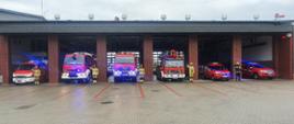 Sześć samochodów pożarniczych stoi w garażu z włączonymi światłami alarmowymi. Garaż ma otwarte bramy wjazdowe. obok samochodów stoją strażacy i salutują. 