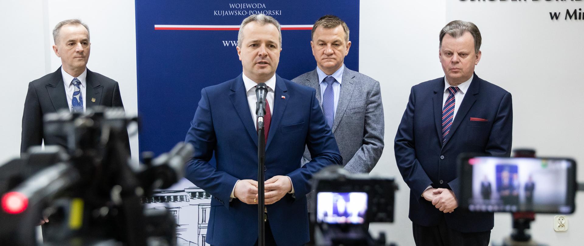 Jeden mężczyzna stojący przed mikrofonem i wypowiadający się, za nim trzech mężczyzn