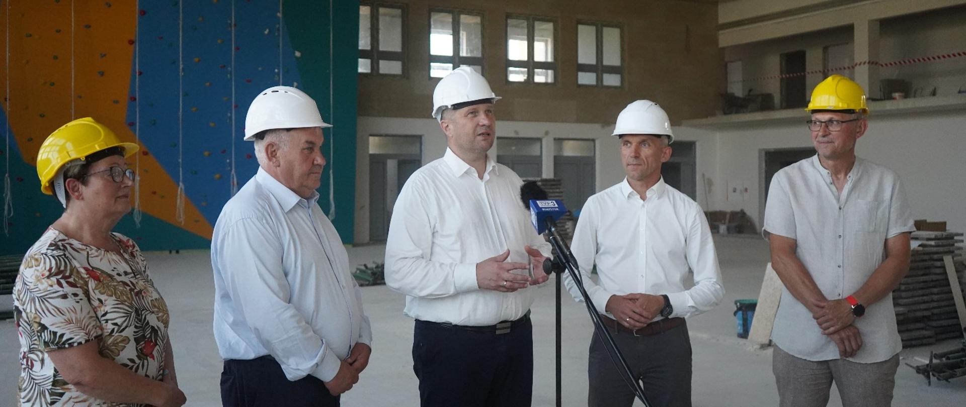 Czterech mężczyzn i kobieta w kaskach budowlanych stoi przed mikrofonem TV w nowo wybudowanej hali sportowej. 