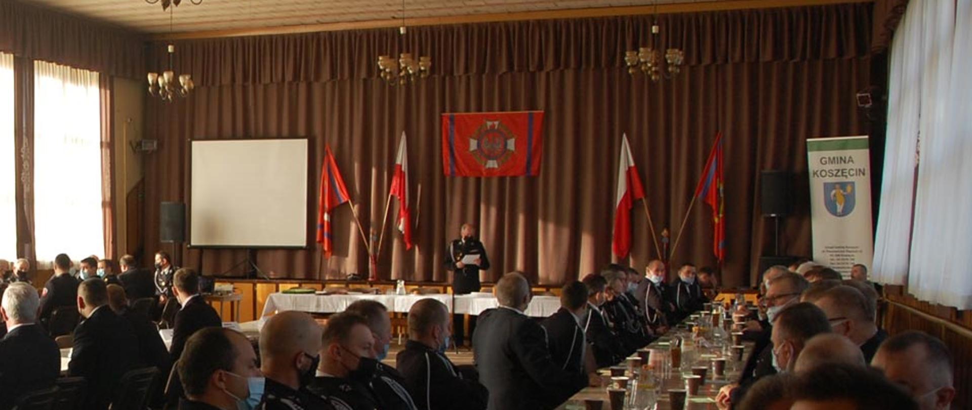 Widok z prawej strony sali bankietowej Ochotniczej Straży Pożarnej w Koszęcinie. Trzy rzędy stołów za którymi siedzą druhowie ubrani w mundury wyjściowe. W tle stół prezydialny za którym stoi prowadzący zebranie. Na ścianie flaga związku ochotniczych straży pożarnych rzeczypospolitej polskiej oraz na drzewcu dwie flagi związku osp i dwie flagi narodowe. Po prawej baner gminy Koszęcin.