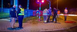 Przejazd drogowo-kolejowy w Gołaszynie, a na nim strażacy i służby kolejowe. W tle rozstawiony czrwony parawan ochronny. Pora nocna.