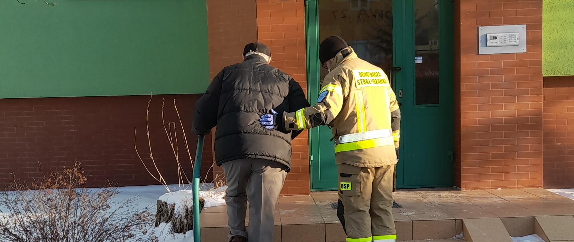 Zdjęcie przedstawia strażaka pomagającego starszej osobie wejść po schodach klatki schodowej. W tle widoczny jest wielorodzinny budynek mieszkalny oraz ośnieżony przed nim chodnik.