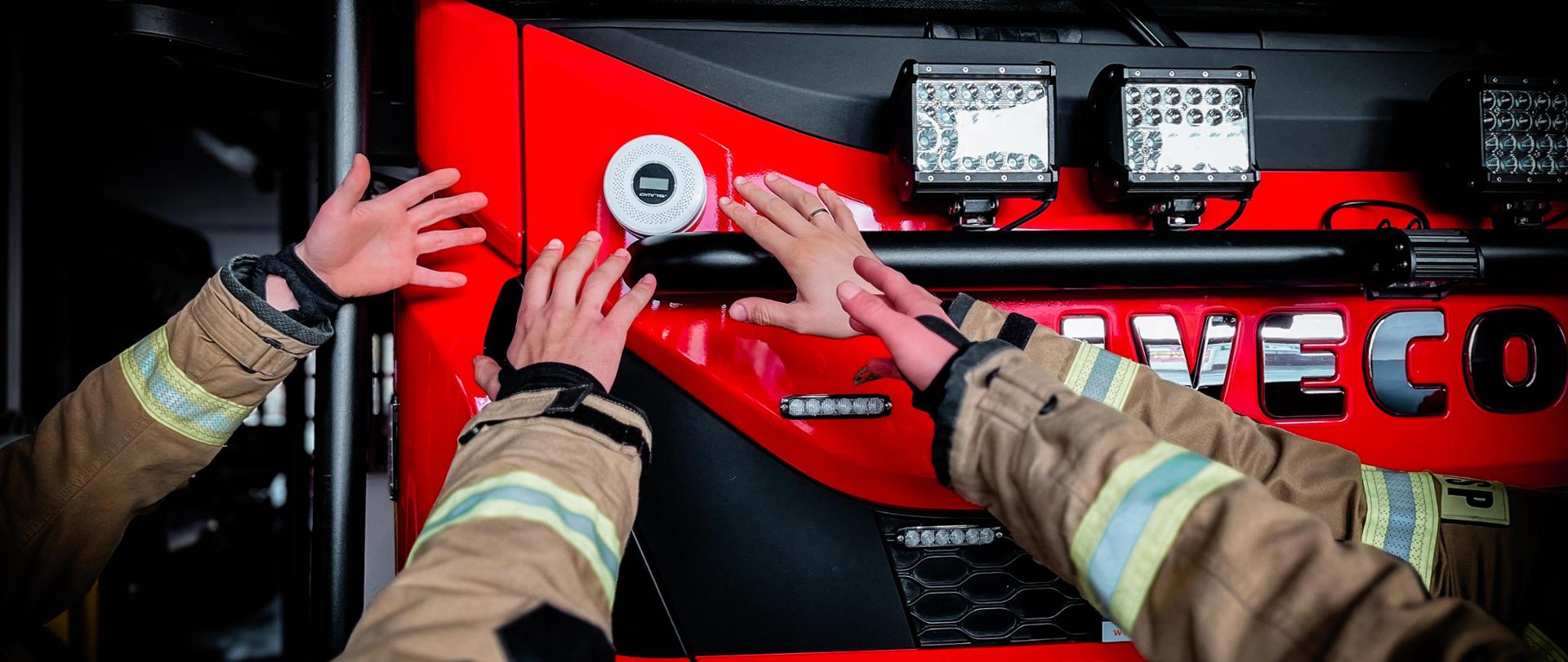 Czujka tlenku węgla położona na wozie strażackim. Po bokach ręce strażaków.