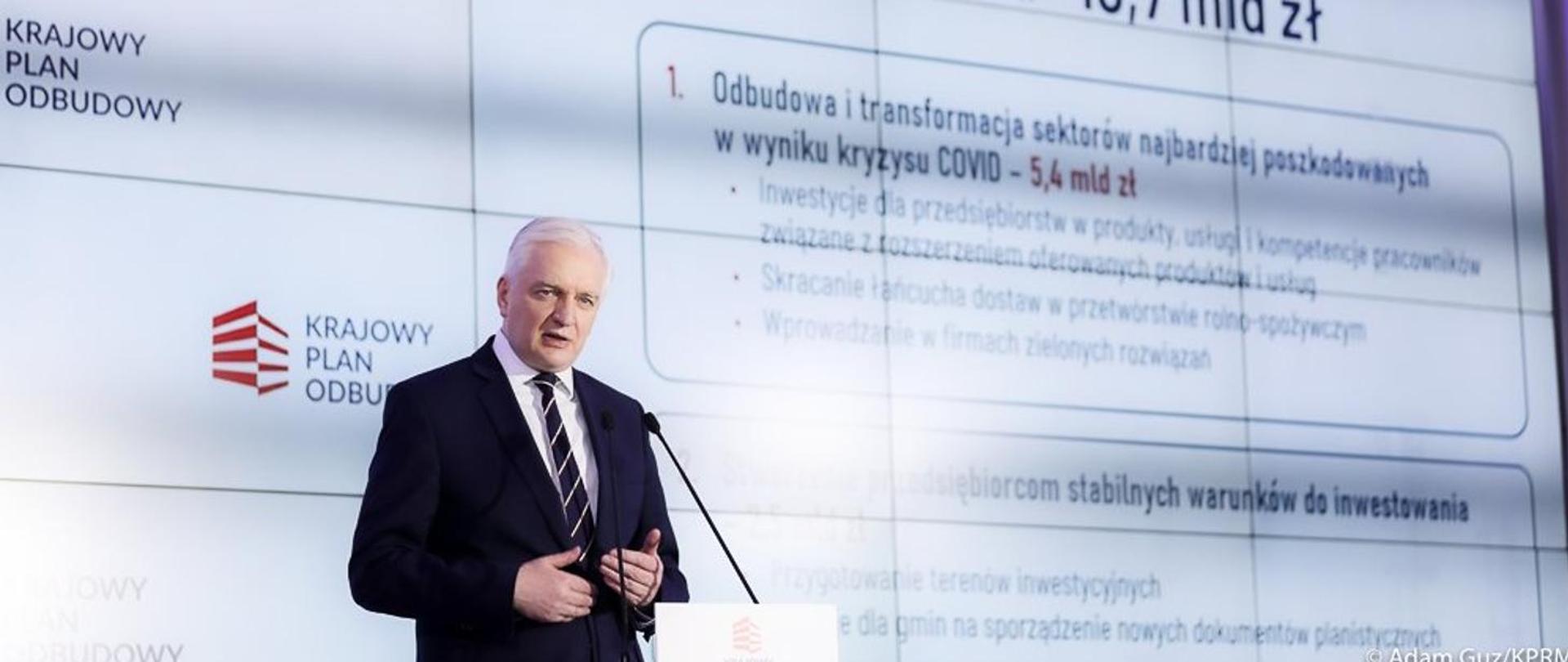 Jarosław Gowin behind the lectern. Behind him, the logo of the National Reconstruction Plan displayed on the big screen.