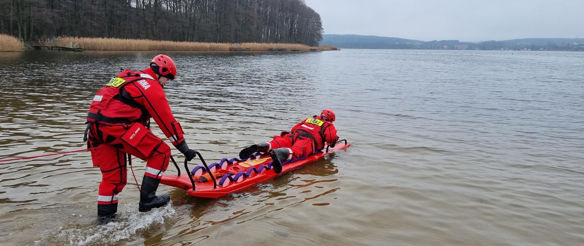 Zdjęcie przedstawia strażaków ćwiczących ratownictwo wodno – lodowe na jeziorze.
W tle jezioro o las.
