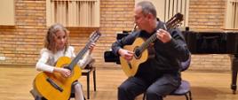 Zdjęcie - z lewej strony siedząca na krześle dziewczynka grająca na gitarze, z prawej nauczyciel z gitarą