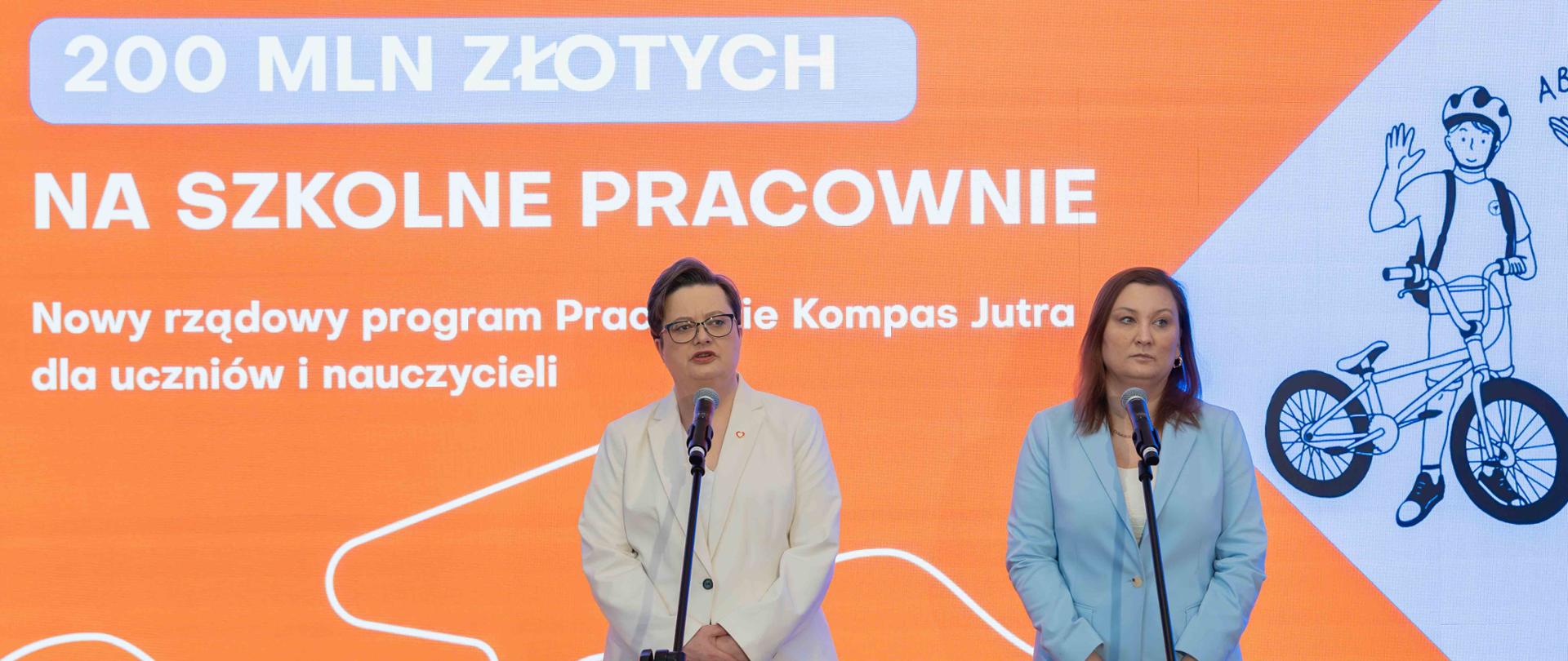 Dwie kobiety (od lewej wiceminister Katarzyna Lubnauer i wiceminister Paulina Piechna-Więckiewicz) w eleganckich garniturach stoją przy mikrofonach na tle pomarańczowego ekranu z napisem „200 MLN ZŁOTYCH NA SZKOLNE PRACOWNIE. Nowy rządowy program Pracownie Kompas Jutra dla uczniów i nauczycieli”.
