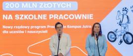 Dwie kobiety (od lewej wiceminister Katarzyna Lubnauer i wiceminister Paulina Piechna-Więckiewicz) w eleganckich garniturach stoją przy mikrofonach na tle pomarańczowego ekranu z napisem „200 MLN ZŁOTYCH NA SZKOLNE PRACOWNIE. Nowy rządowy program Pracownie Kompas Jutra dla uczniów i nauczycieli”.