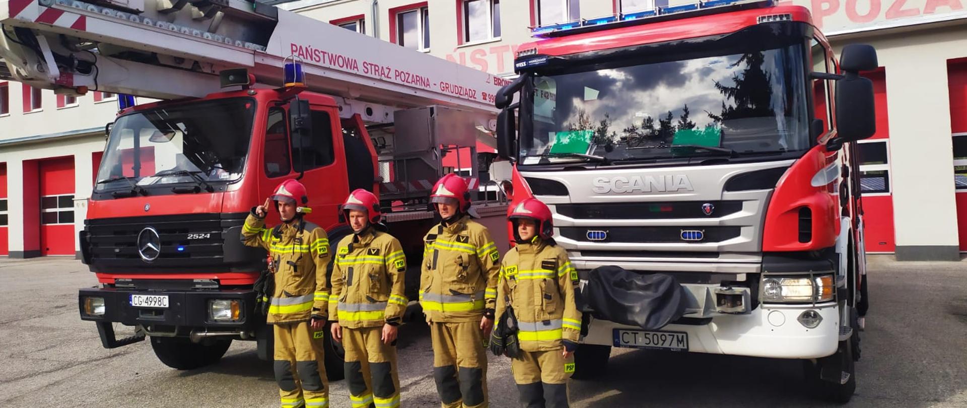 Zdjęcie przedstawia 2 czerwone, ciężarowe samochody strażackie stojące przed budynkiem jednostki. Przed nimi w rzędzie stoi 4 strażaków ubranych w piaskowe mundury specjalne i czerwone hełmy . Dowódca oddaje honor.