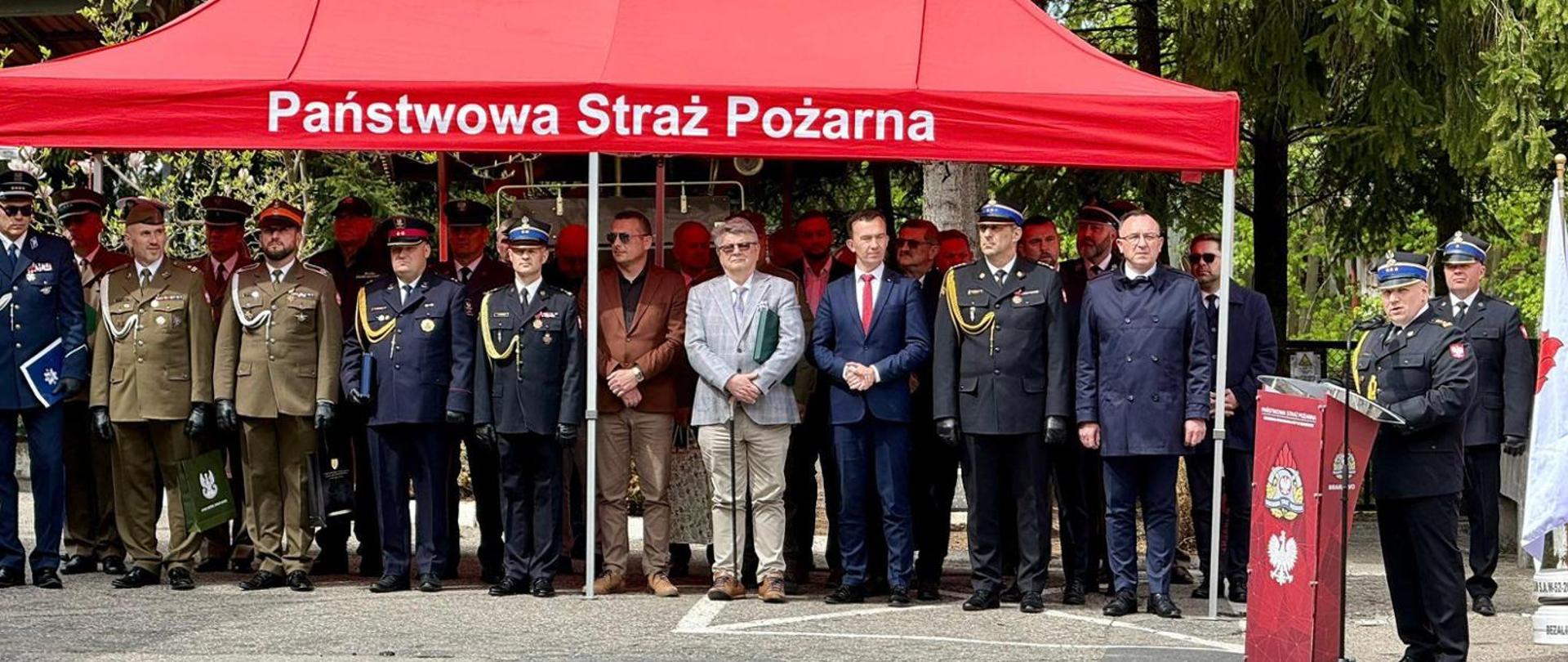 Na zdjęciu pod czerwonym namiotem z napisem PSP, zaproszeni goście podczas obchodów Dnia Strażaka w Braniewie, przemawia komendant z Braniewa