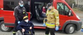 Przy zaparkowanym samochodzie strażackim typu bus koloru czerwonego dwóch strażaków w mundurach wiezie starszą kobietę na wózku inwalidzkim. Samochód ma otwarte tylne drzwi boczne. Przez drzwi widać siedzącą w samochodzie kolejną kobietę w maseczce ochronnej.