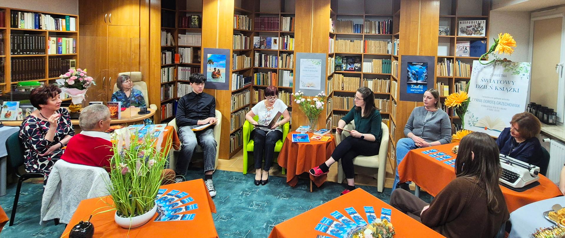 W pomieszczeniu biblioteki, na tle ściany z regałami wypełnionymi książkami w fotelach siedzi dziewięć osób w różnym wieku. Między nimi stoją stoliki nakryte pomarańczowymi obrusami i udekorowane kompozycjami roślinnymi, książkami i niebieskimi zakładkami do książek. Po prawej stronie na stoliku stoi maszyna do pisania, z za nią plakat informujący o światowym dniu książki i spotkaniu autorskim z panią Dorotą Grzechowiak.