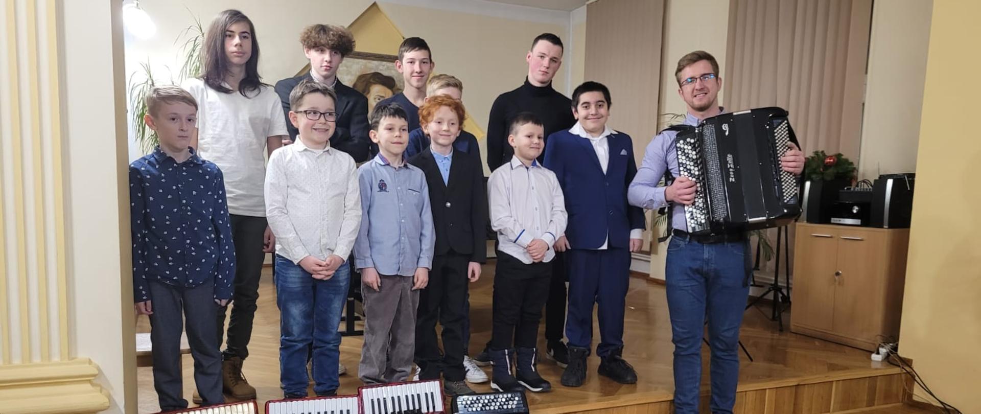 Na zdjęciu stojąca grupa uczniów wraz nauczycielem Tomaszem Brusińskim, przed nimi na pierwszym planie leżące akordeony. Zdjęcie wykonane w auli PSM I st. w Kłodzku podczas koncertu "Akordeonowe Music Party" - 20 stycznia 2023