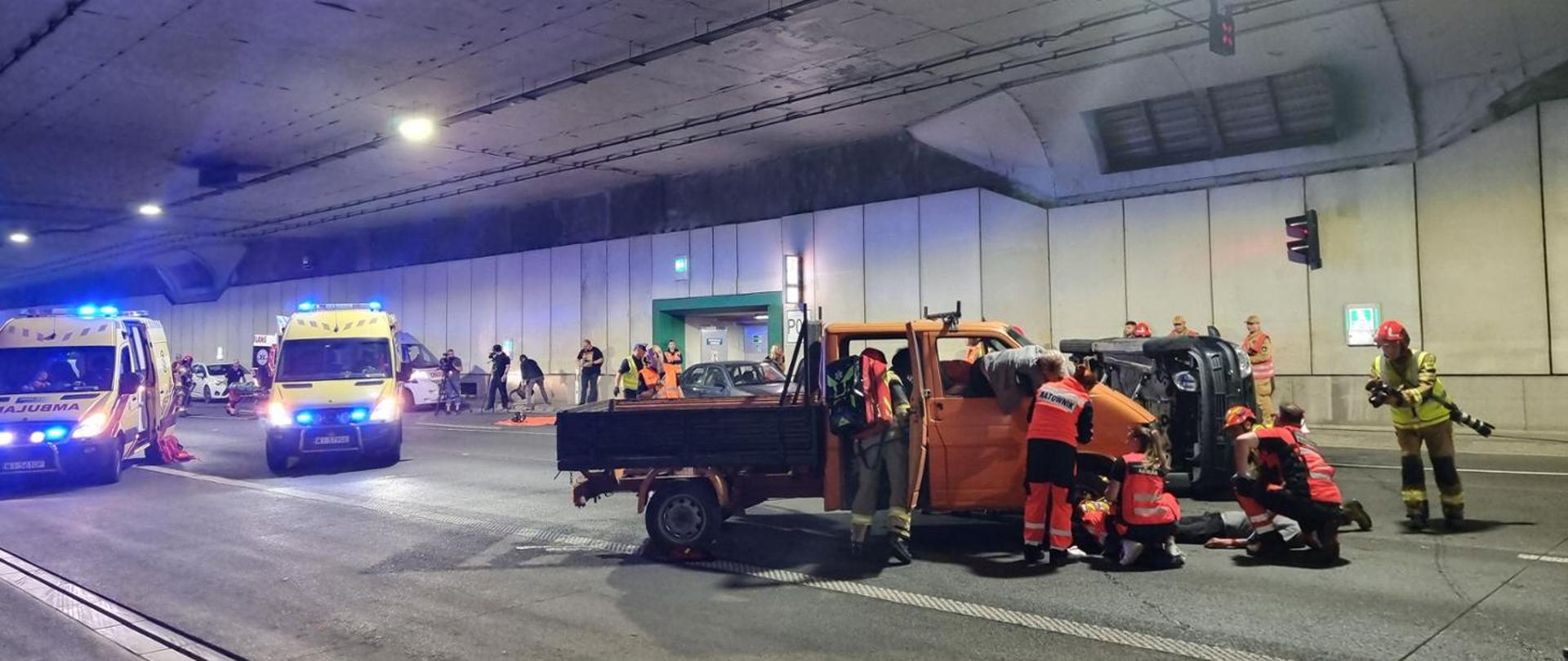 Ćwiczenia służb ratunkowych w tunelu drogowym. Na jezdni znajduje się przewrócony na bok samochód osobowy oraz pomarańczowy samochód dostawczy. Ratownicy medyczni i strażacy prowadzą działania przy poszkodowanych, a w tle stoją karetki pogotowia z włączonymi sygnałami świetlnymi. W tunelu widoczne są także inne pojazdy służb oraz osoby uczestniczące w ćwiczeniach. Cała scena przedstawia symulację wypadku drogowego i skoordynowaną akcję ratunkową.