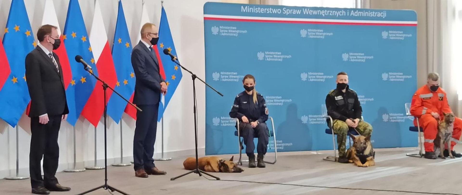 zdjęcie z konferencji, na którym widać stojących ministra swia, prowadzącego konferencję, oraz policjantkę strażnika granicznego i strażaka z psami służbowymi. Funkcjonariusze siedzą na krzesłach w tle flagi polski i unii europejskiej i baner ministerstwa swia