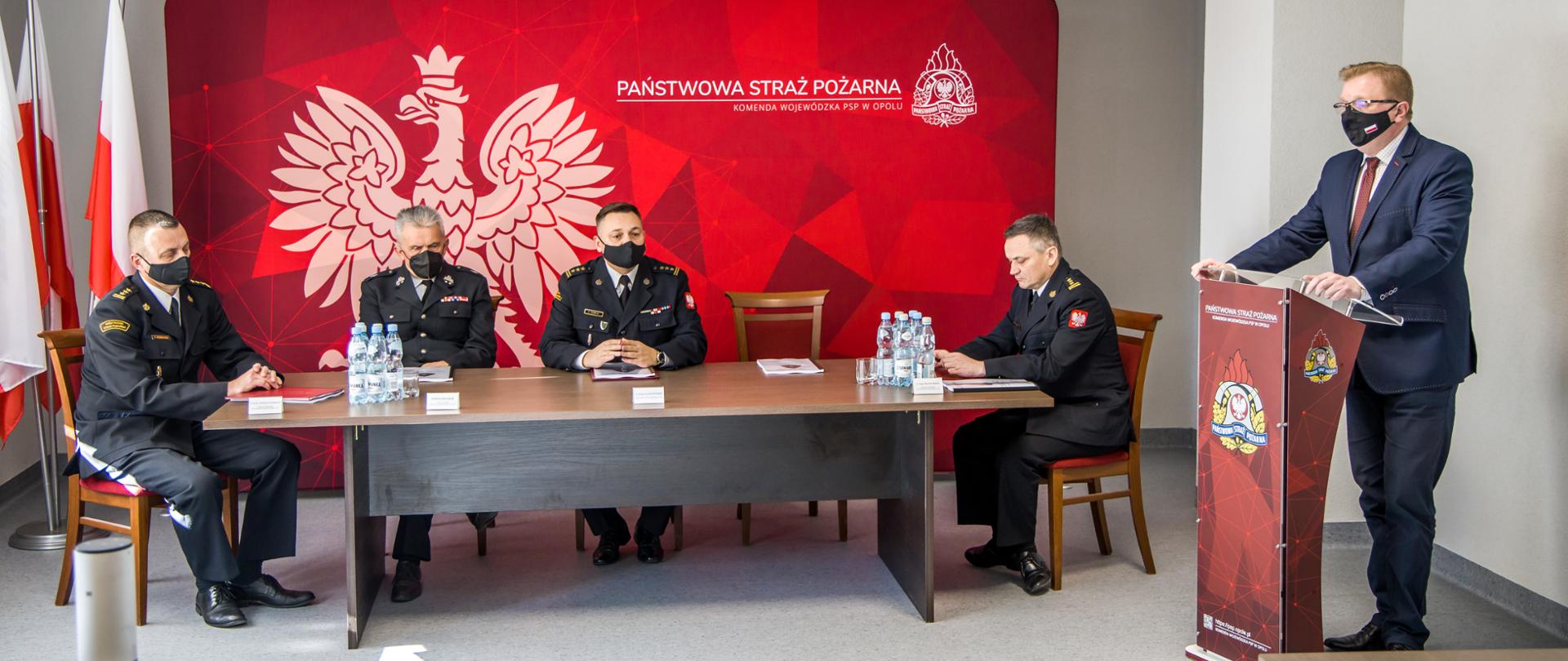 Odprawa z kadrą kierowniczą PSP Opole. Na zdjęciu 4 mężczyzn zasiadających za stołem, obok - po prawej stronie kadru, mównica za którą stoi i przemawia wicewojewoda opolski 