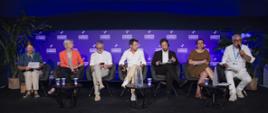 Na zdjęciu widać panel dyskusyjny odbywający się na scenie podczas konferencji. Siedem osób zajmuje miejsca w fotelach ustawionych w jednym rzędzie, a przed nimi znajdują się niewielkie stoliki z wodą i kubkami. Osoba siedząca po prawej stronie trzyma mikrofon i prawdopodobnie zabiera głos w trakcie rozmowy. Tło sceny stanowi podświetlana na fioletowo ściana z powtarzającym się napisem „Les Rencontres Économiques Aix-en-Provence”. Po obu stronach sceny ustawiono duże rośliny doniczkowe, które dekorują przestrzeń. Całość jest profesjonalnie oświetlona, a uczestnicy panelu mają przed sobą notatki lub dokumenty.