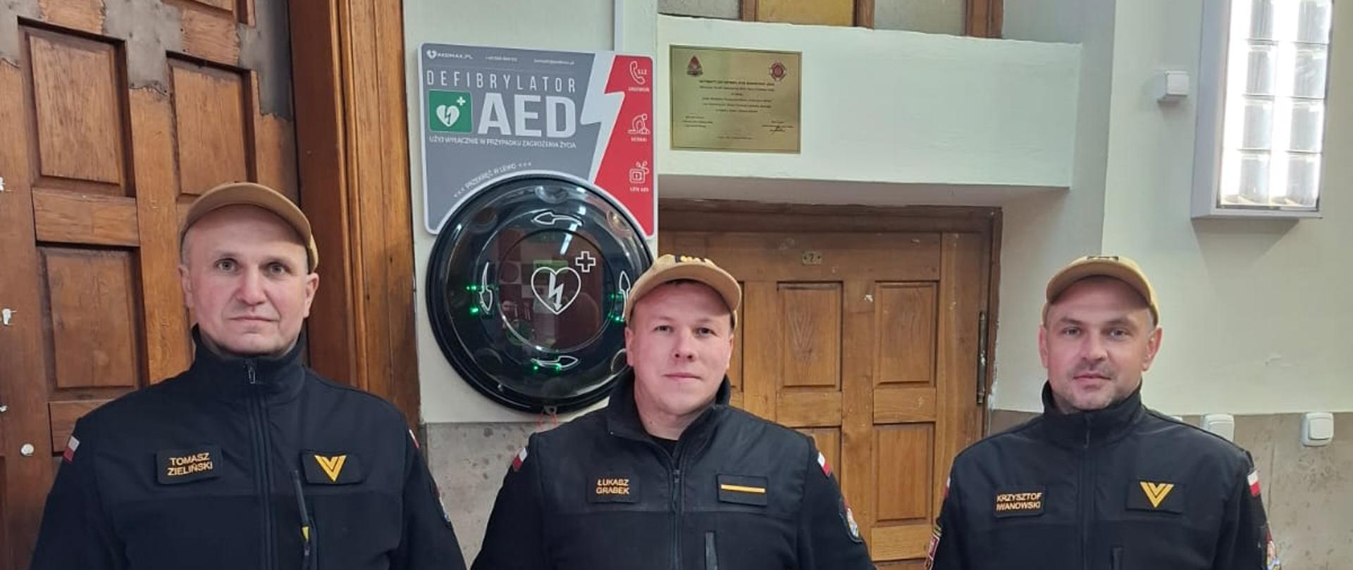 Trzech strażaków stojących w przedsionku kościoła. Strażacy stoją na tle nowo zamontowanego defibrylatora. 