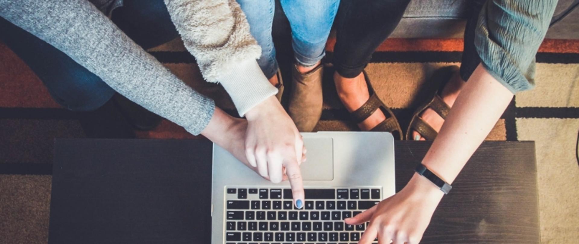 Zbliżenie na dłonie trzech osób wskazujących na klawiaturę laptopa; scena sugeruje współpracę lub dyskusję.