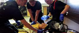 Zdjęcie zrobione w sali szkoleniowej podczas kursu kwalifikowanej pierwszej pomocy. Trzech strażaków ćwiczy udzielanie pierwszej pomocy na fantomie podłączonym defibrylatora. 