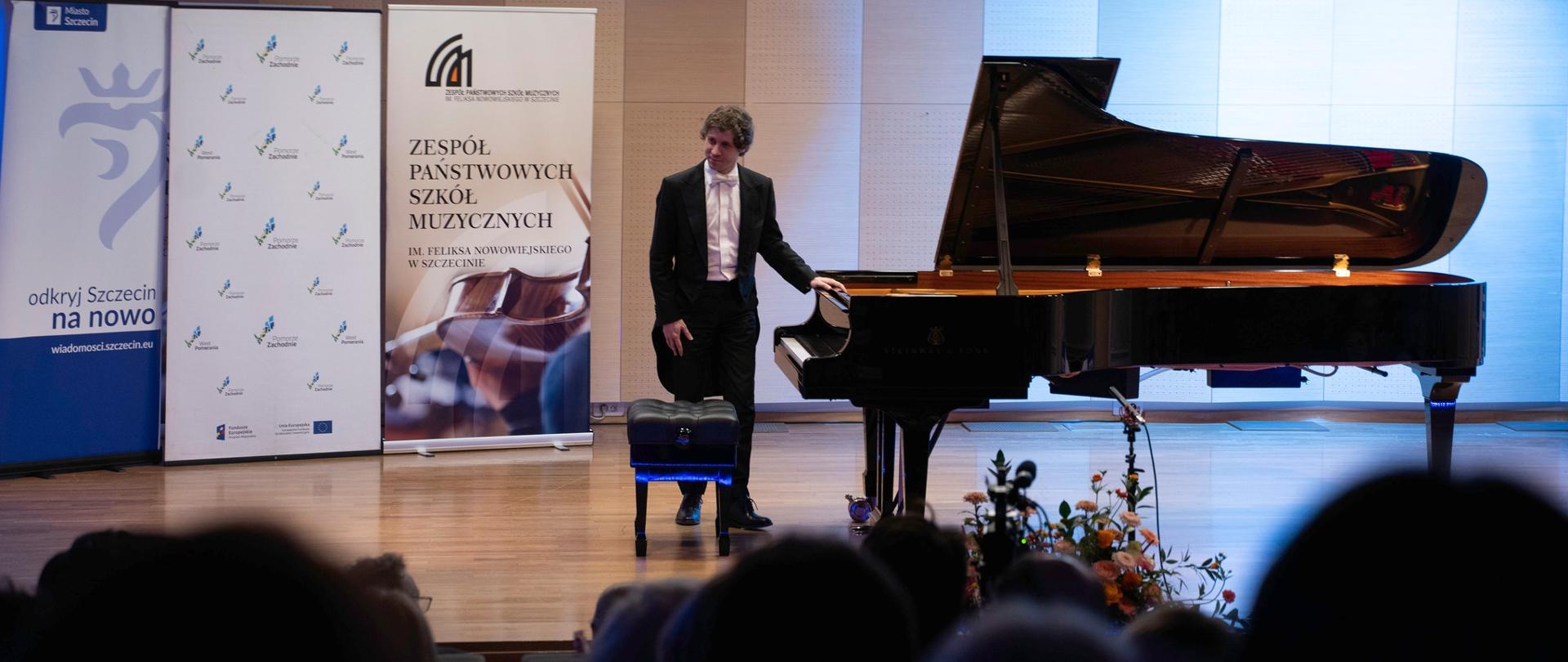 zdjęcie przedstawia pianistę kłaniającego się przy fortepianie na scenie sali koncertowej przed rozpoczęciem koncertu, w tle banery szkoły i sponsorów