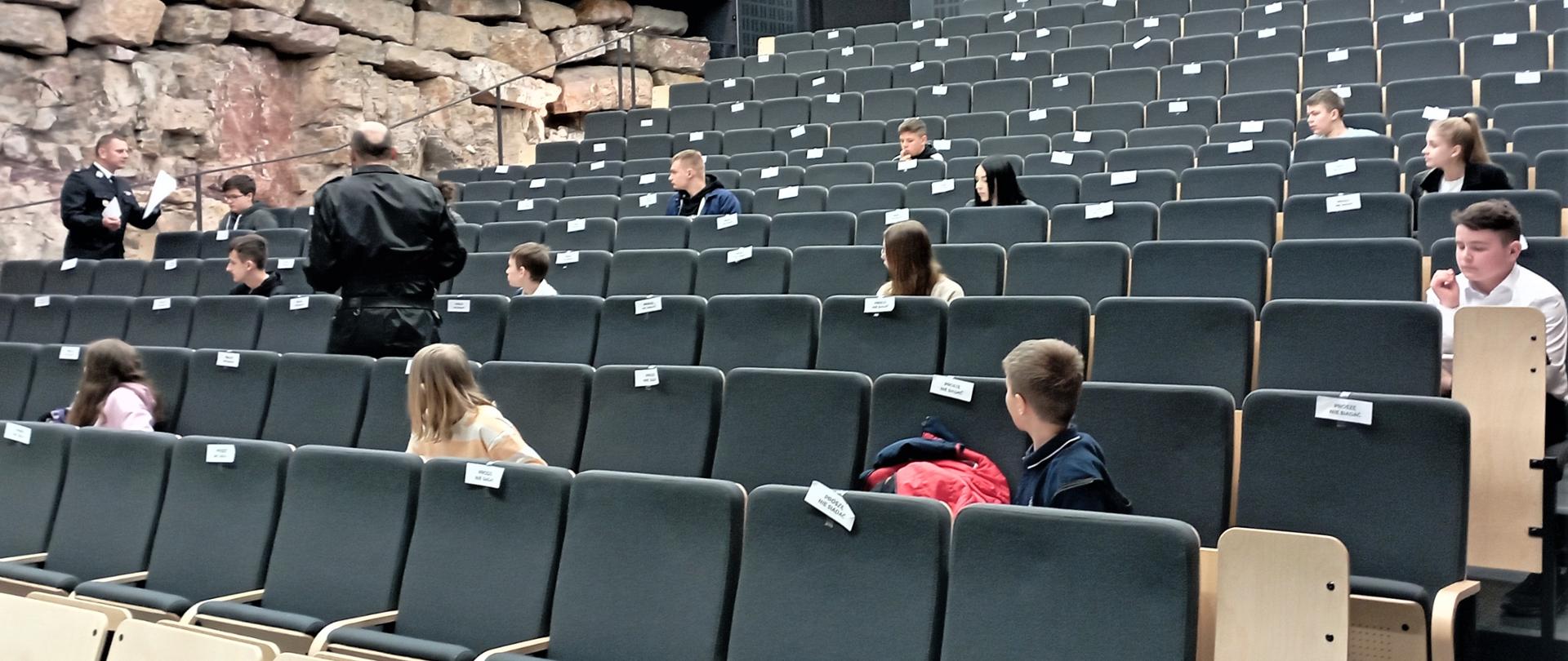 Na zdjęciu przedstawiono uczestników Ogólnopolskiego Turnieju Wieży Pożarniczej w jednej z gmin powiatu kieleckiego. Zawodnicy siedzą w pięknej auli a członkowie jury wyjaśniają sposób wypełniania testu.