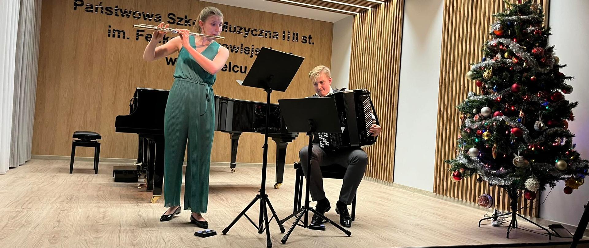 Flecistka i akordeonista podczas występu na scenie w auli koncertowej, z prawej strony udekorowana choinka. 