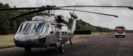 Na pierwszym planie, stoi na asfaltowym pasie, szary śmigłowiec Black Hawk z napisem POLICJA, , w tle czerwony wóz strażacki.
