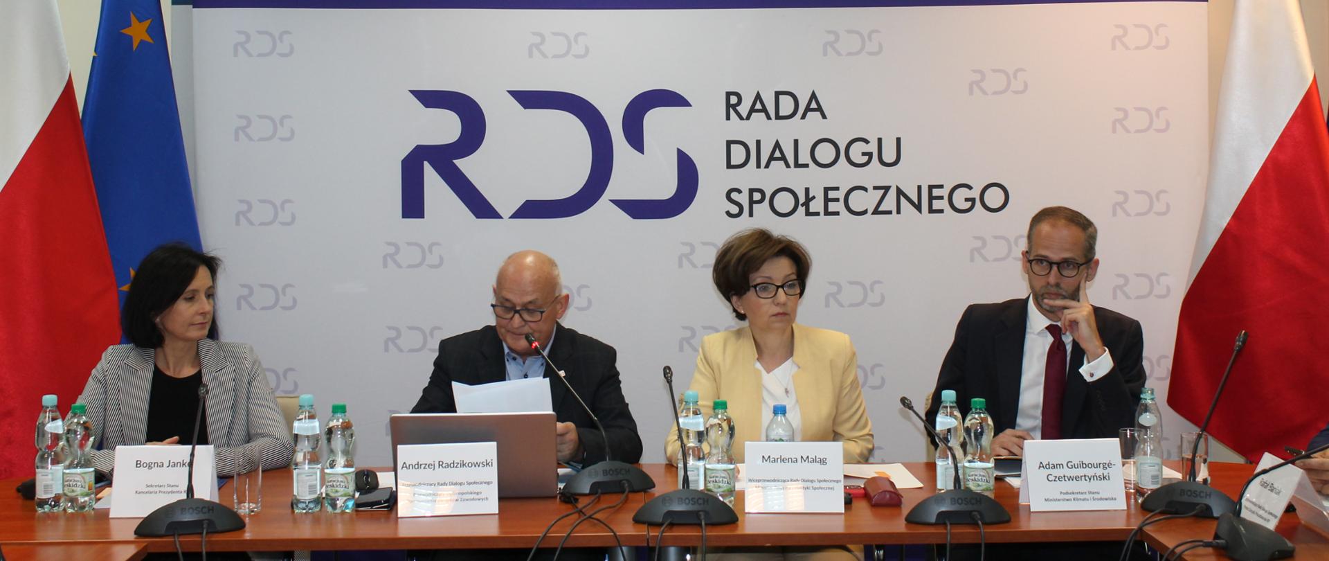 Zdjęcie przedstawia siedzące za stołem prezydialnym cztery osoby. Licząc od lewej strony: Bogna Janke – Sekretarz Stanu w Kancelarii Prezydenta, Andrzej Radzikowski - Przewodniczący Rady Dialogu Społecznego, Marlena Maląg - Wiceprzewodnicząca Rady, Minister Rodziny i Polityki Społecznej oraz Adam Guibourgé-Czetwertyński – Podsekretarz Stanu w Ministerstwie Klimatu i Środowiska. W tle osób baner z napisem Rada Dialogu Społecznego. Po lewej stronie dwie flagi: Polski i UE a po prawej stronie jedna flaga Polski. Nad banerem wisi godło Polski.