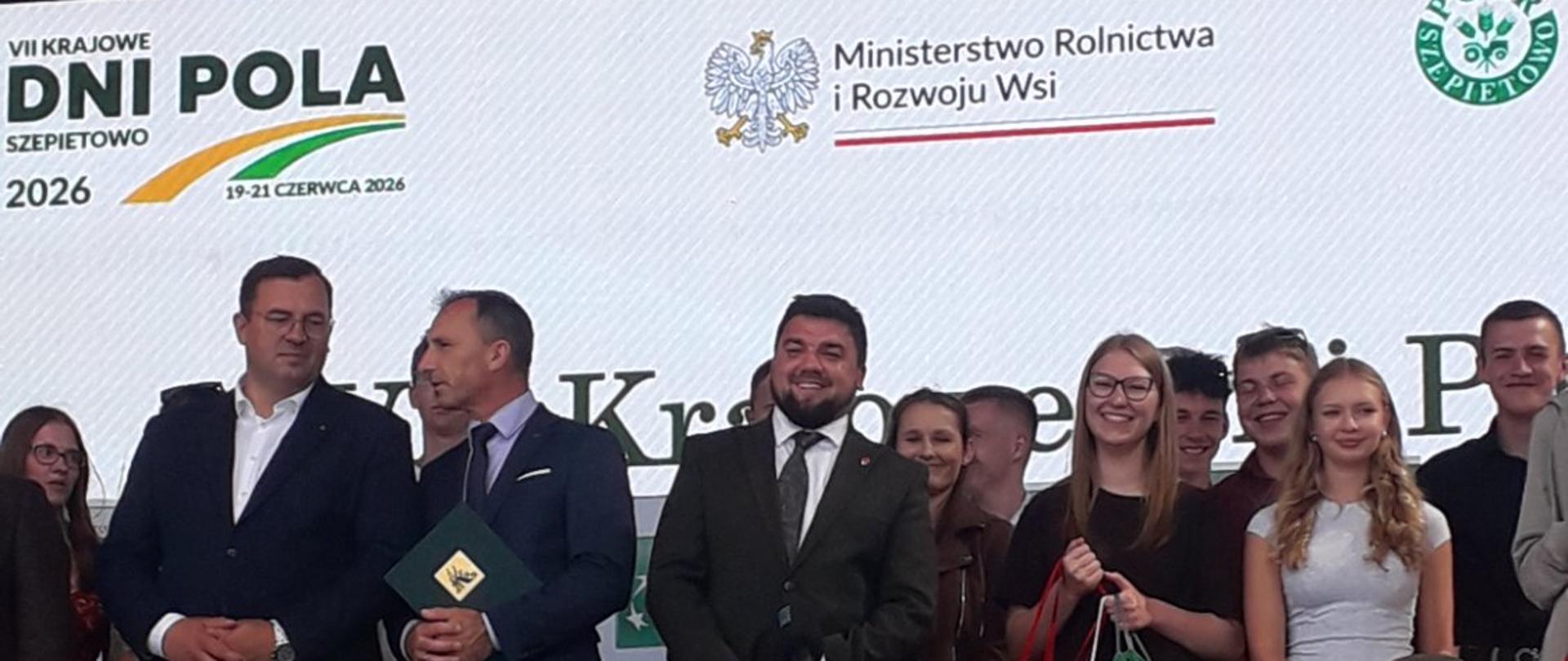Grupa osób stoi na udekorowanej kwiatami scenie podczas wydarzenia rolniczego. W tle duży baner z napisem ‘VII Krajowe Dni Pola Szepietowo 2026, 19–21 czerwca’ oraz logotypami Ministerstwa Rolnictwa i PODR Szepietowo.