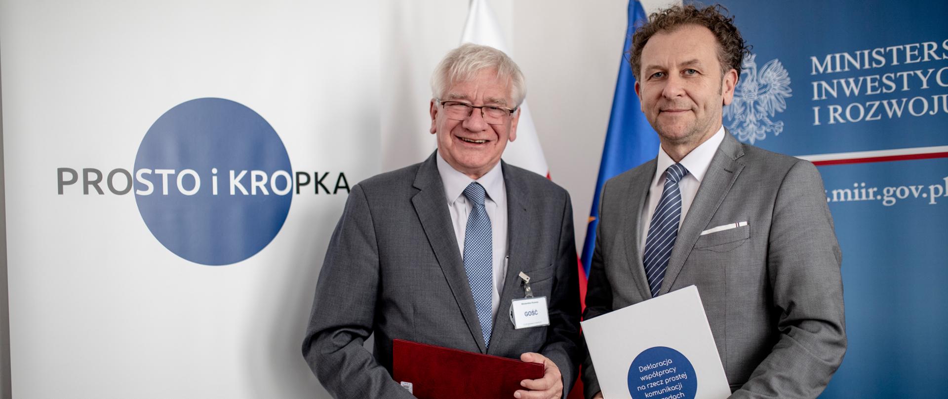 Dwóch mężczyzna trzyma w ręku podpisane umowy, stoją przed ściankami na jednej jest napisane "prosto i kropka" na drugim "Ministerstwo Inwestycji i Rozwoju"