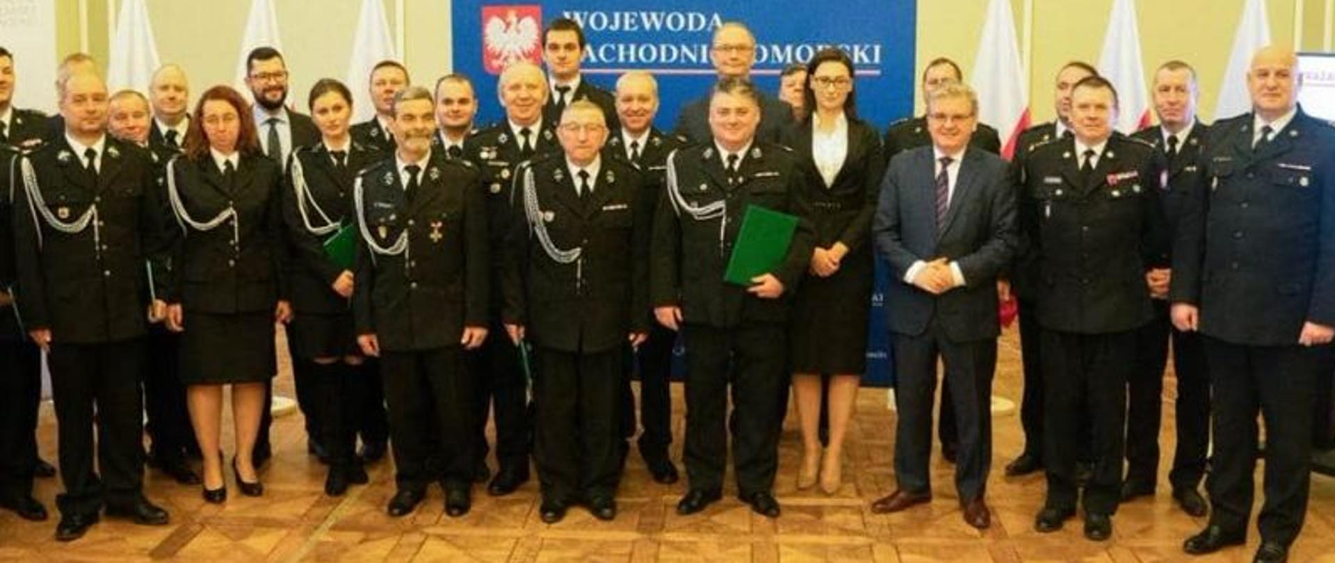 Zdjęcie grupowe wykonane wewnątrz budynku na sali gdzie przeprowadzono wręczenie dotacji. Strażacy stoją w grupie, w mundurach wyjściowych. Wśród strażaków stoją urzędnicy, posłowie.