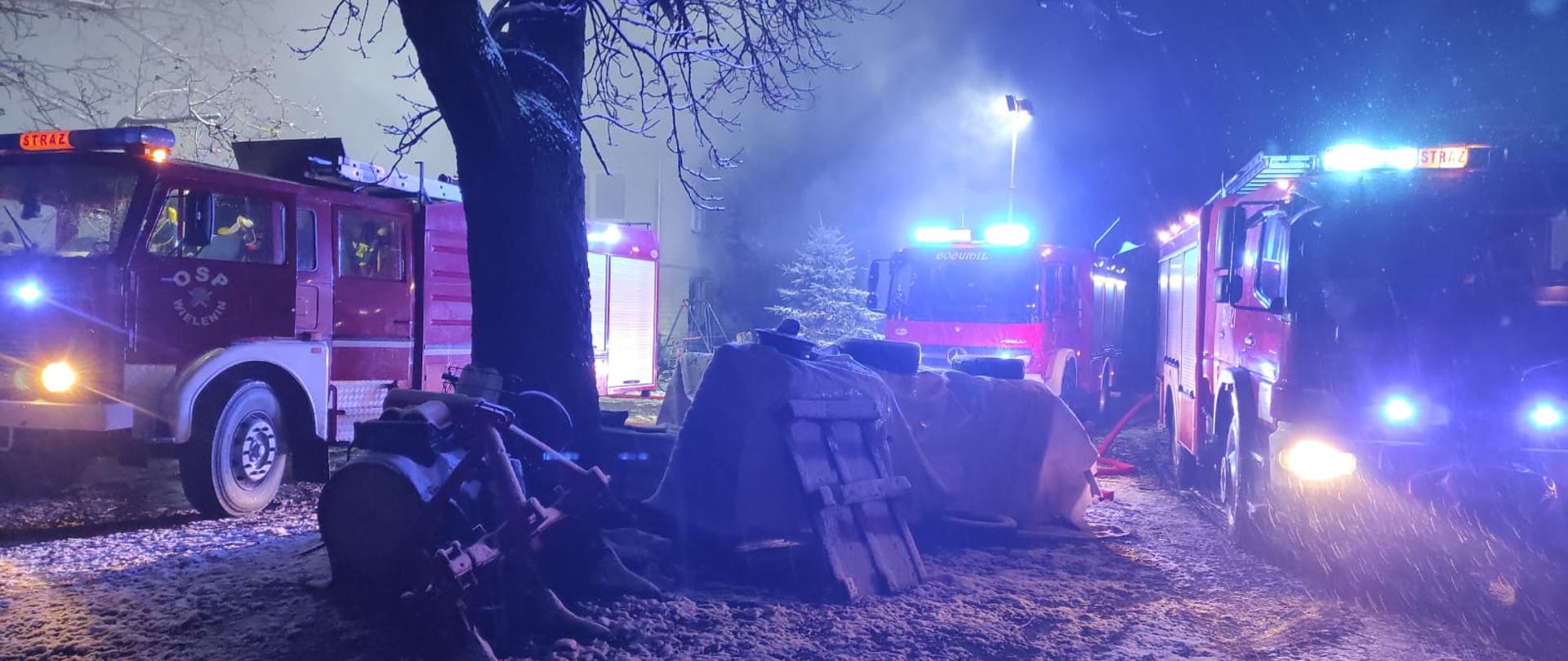 Zdjęcie przedstawia trzy wozy straży pożarnej z włączonymi światłami ostrzegawczymi stojące na posesji na której powstał pożar