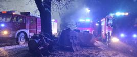 Zdjęcie przedstawia trzy wozy straży pożarnej z włączonymi światłami ostrzegawczymi stojące na posesji na której powstał pożar