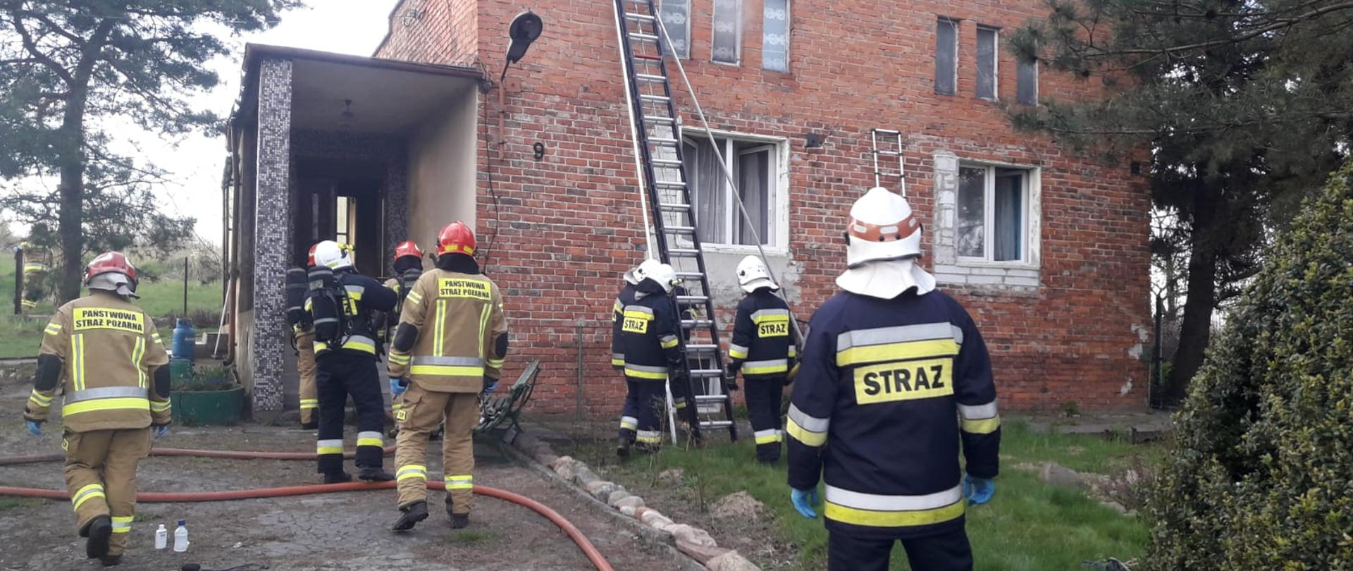 Na zdjęciu widać budynek parterowy, z czerwonej cegły, z płaskim dachem. Z okien poddasz wydobywa się dym. Przed budynkiem widać strażaków w ubraniach specjalnych i hełmach. Pomiędzy nimi widoczna czerwona linia wężowa. O ścianę frontową budynku oparta czarna drabina D10W. Po prawej i lewej stronie zdjęcia widać drzewa i krzewy. Na dachu budynku antena satelitarna w kolorze białym. 